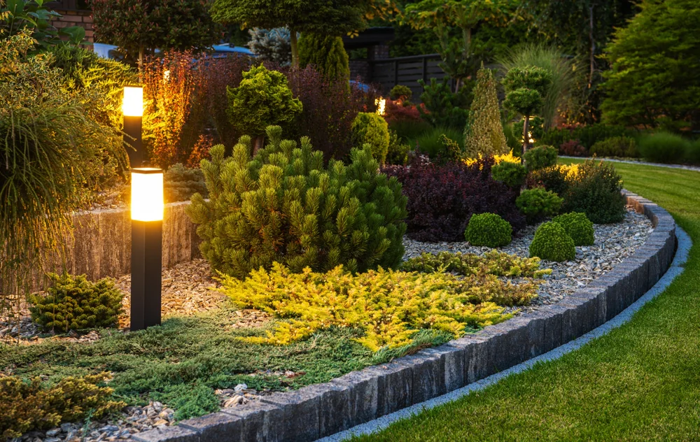 A landscaped garden features a variety of bushes and shrubs bordered by stone edging, with glowing modern pathway lights illuminating the greenery in the evening.