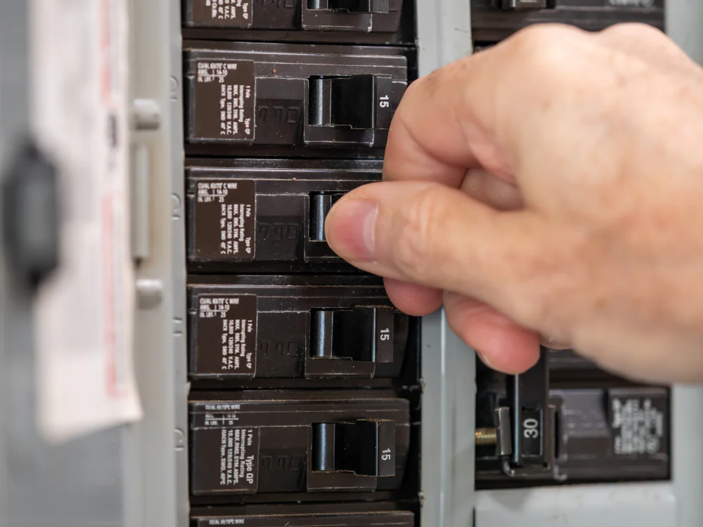 A hand flips a switch on a row of black circuit breakers inside an electrical panel.