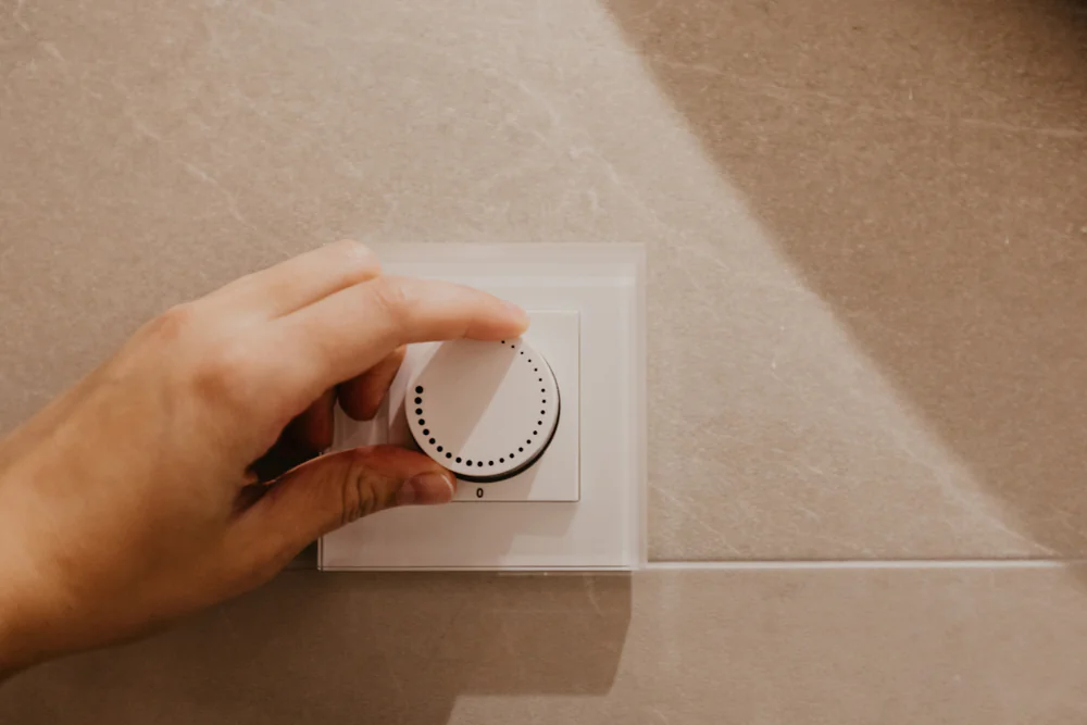 A hand is adjusting a circular thermostat dial mounted on a beige tiled wall.