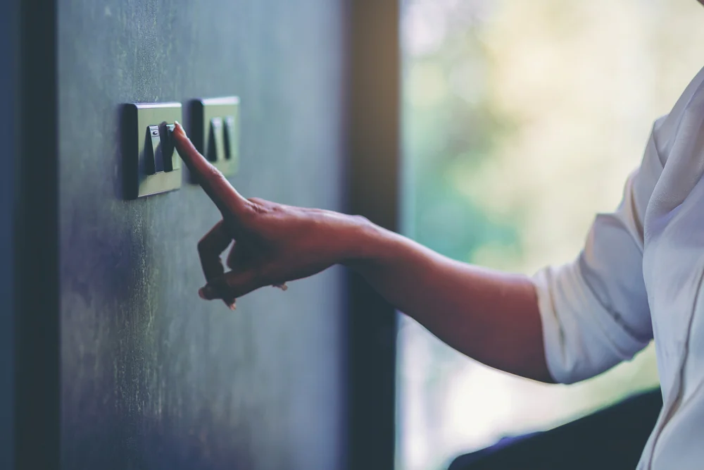 A person wearing a white shirt is pressing a light switch on a dark-colored wall, with their arm extended and finger touching the switch.