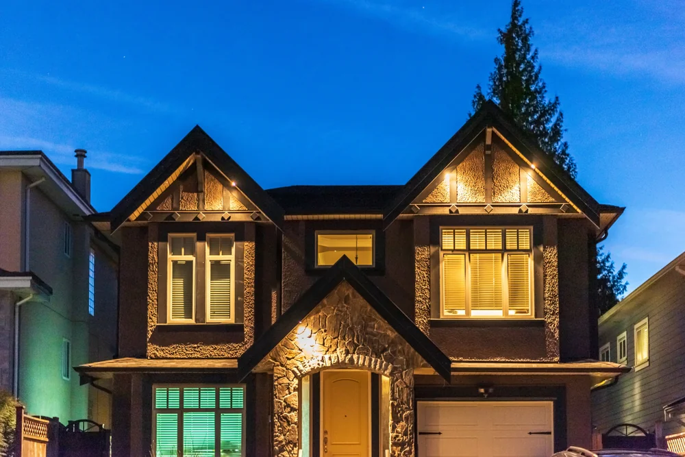 A two-story house with exterior lights on, stone accents around the entrance, and large windows, photographed at dusk with a deep blue sky in the background.