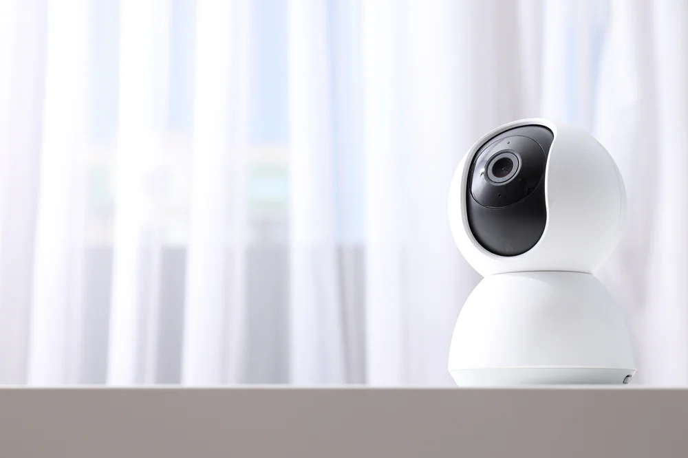A white, dome-shaped security camera sits on a white surface in front of sheer white curtains, with soft natural light filling the scene.