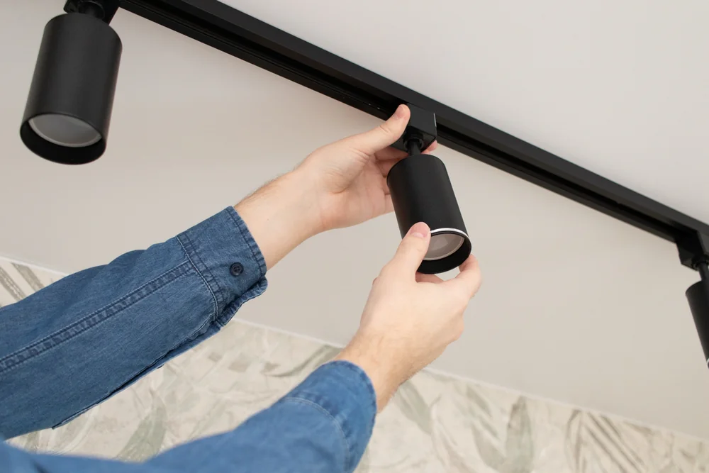 A person wearing a blue shirt adjusts a black track light fixture mounted on a ceiling, with patterned wallpaper visible in the background.