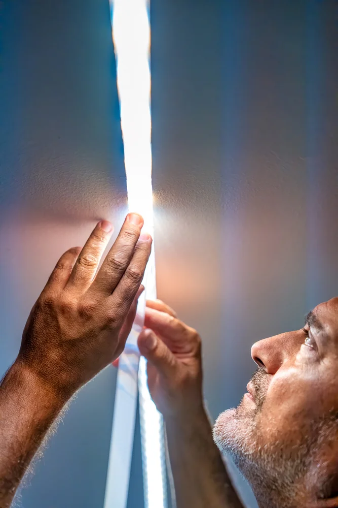 A man closely examines or installs a brightly glowing LED strip light on a wall, with his face and hands illuminated by the intense white light.