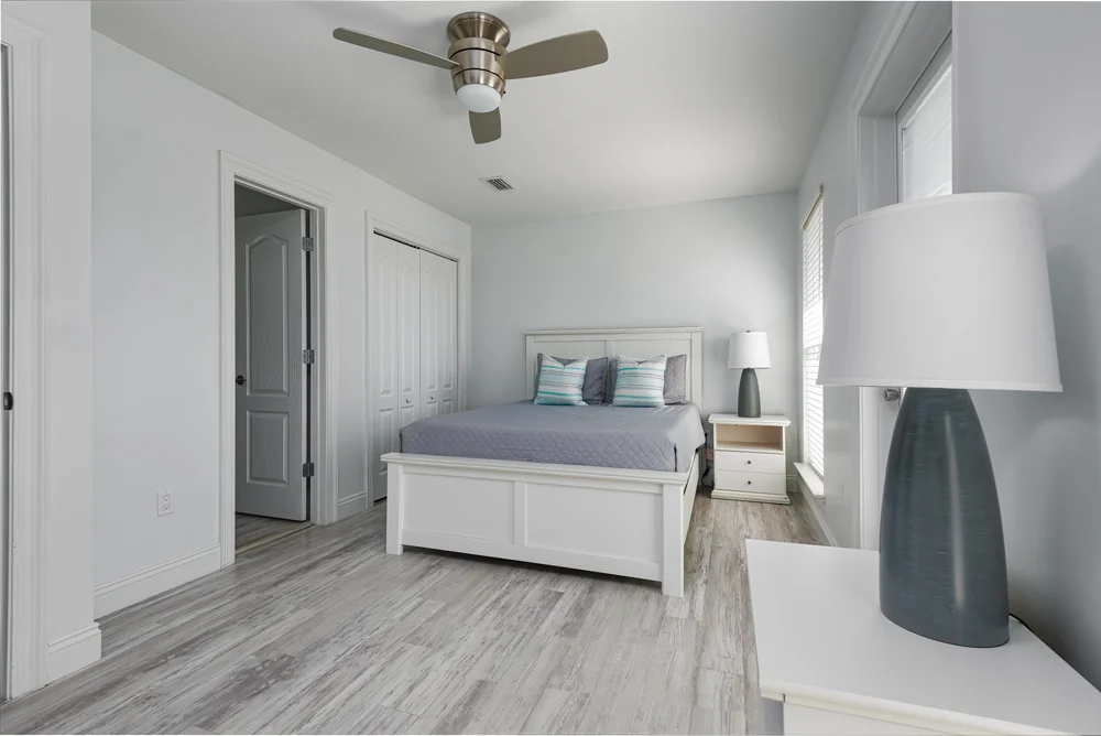 A modern bedroom with white walls and light wood flooring features a white bed with gray bedding, two nightstands with lamps, a ceiling fan, and a closed closet with double doors. Natural light fills the room.