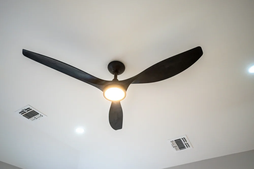 A modern black ceiling fan with three blades and a built-in light is mounted on a white ceiling, with two air vents and recessed lighting also visible nearby.