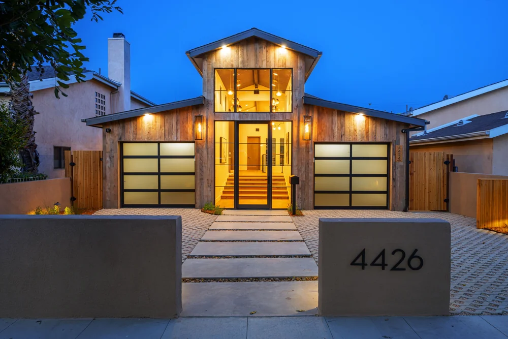 Modern two-story house with wood siding, large glass windows, double frosted glass garage doors, illuminated exterior lights, and the address "4426" on a concrete wall at the entrance, pictured at dusk.