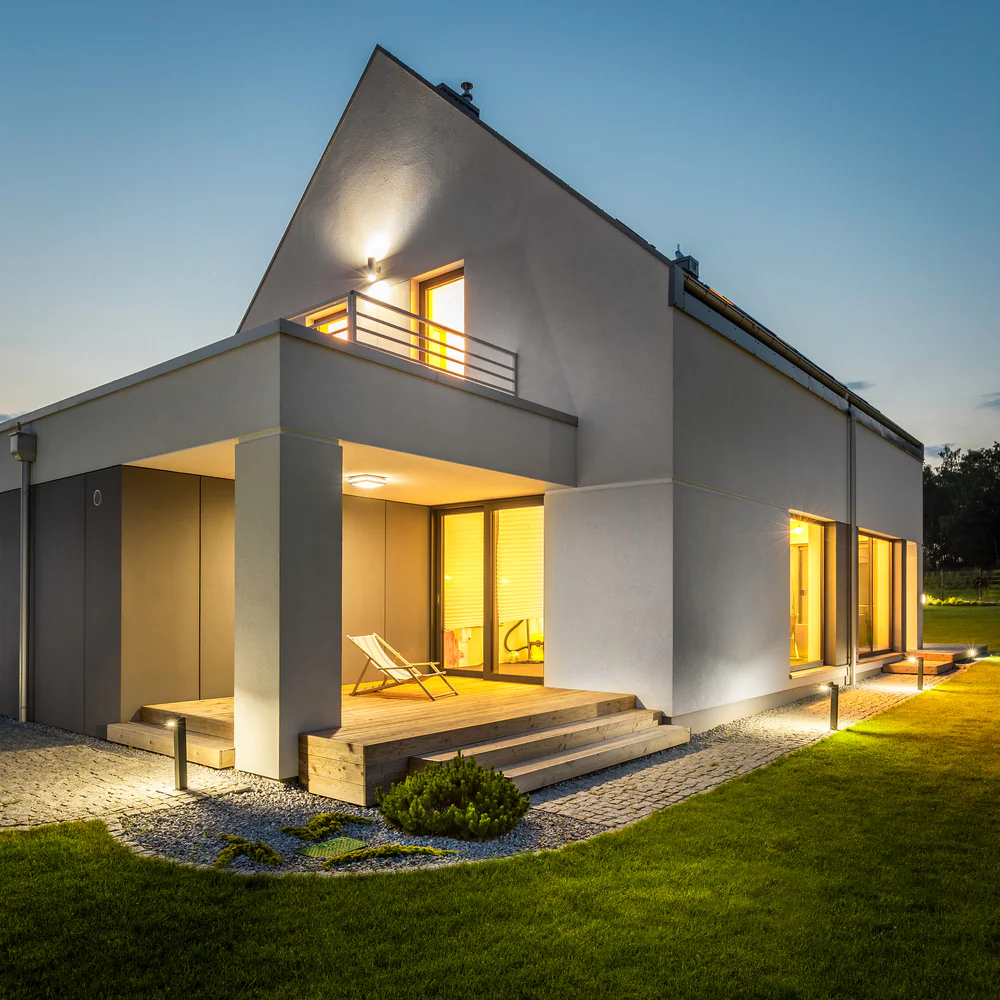 Modern two-story house at dusk with large windows glowing warmly, a wooden porch with a lounge chair, sloped roof, and neatly manicured lawn under a clear evening sky.