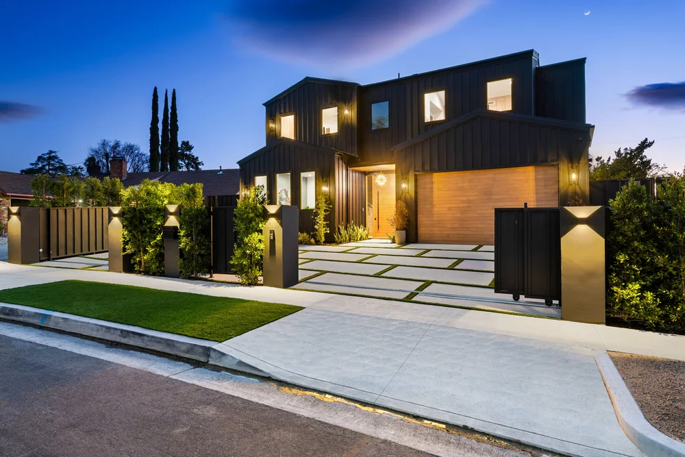 Modern two-story house at dusk with dark exterior, large windows, warm lighting, a wooden accent wall, and a landscaped front yard featuring geometric concrete tiles and greenery behind a sleek fence.