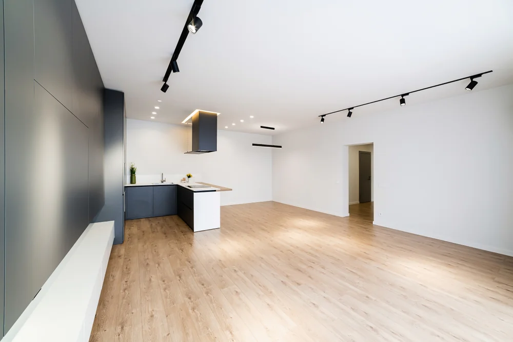 Modern, empty open-plan room with light wood flooring, white walls, black accent wall, and a sleek kitchen area with dark cabinets and an island. Ceiling spotlights and track lighting illuminate the space.