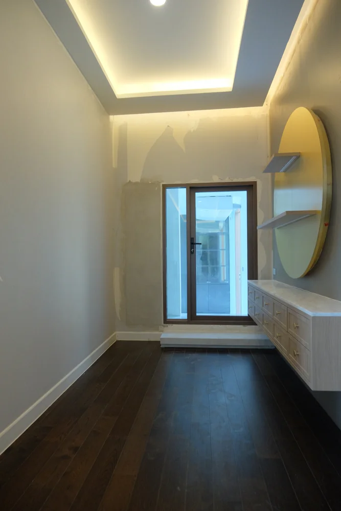 A narrow room with dark wooden floors, light gray walls, a window at the end, built-in shelves, a round mirror, and a floating cabinet beneath it. The ceiling has recessed lighting.