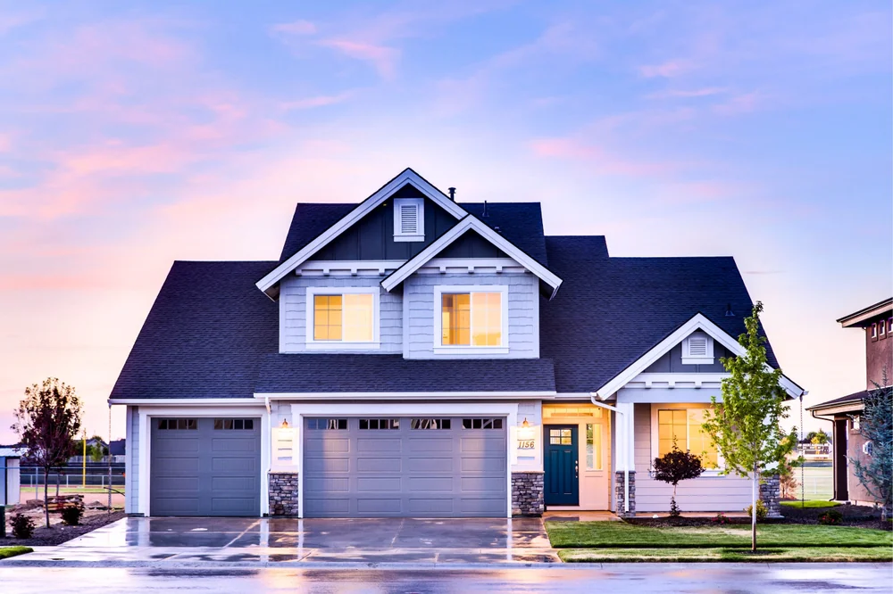 A two-story modern suburban house with gray siding, large windows, and three garage doors, illuminated warmly inside, sits by a wet driveway at sunset with a partly cloudy sky.