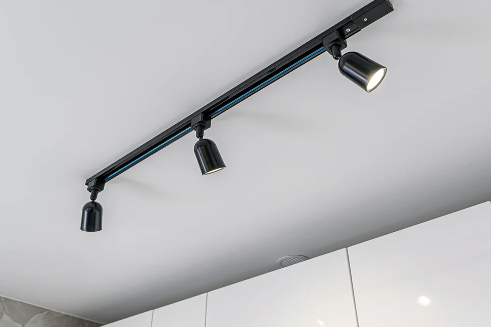 A black three-light track lighting fixture with adjustable spotlights is mounted on a white ceiling above white cabinets. One spotlight is turned on, illuminating the area below.