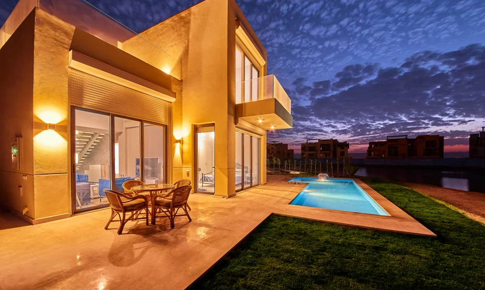 Modern two-story house with glowing exterior lights, swimming pool, and patio seating, set against a dramatic evening sky with scattered clouds.