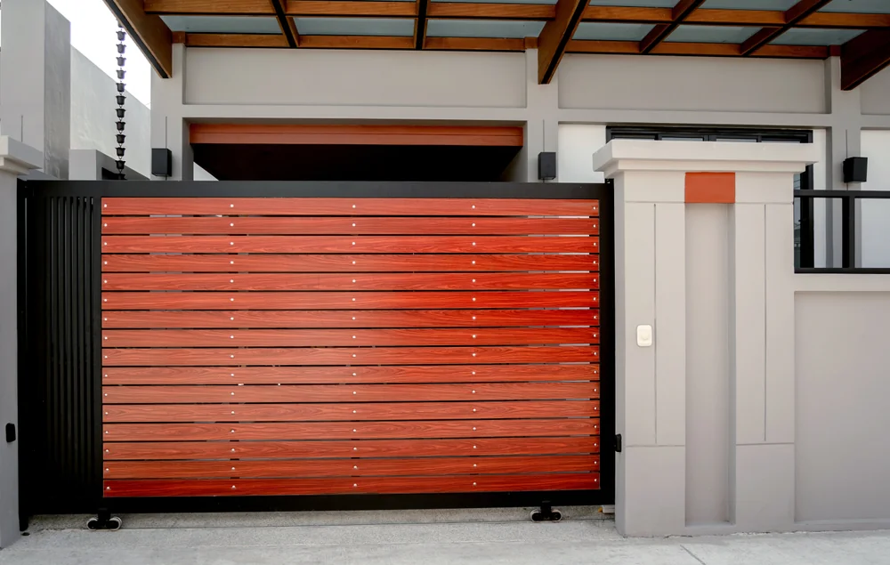 A modern house entrance features a sliding gate made of horizontal wooden planks with a black metal frame, set against a light gray concrete wall and a partially visible carport roof.