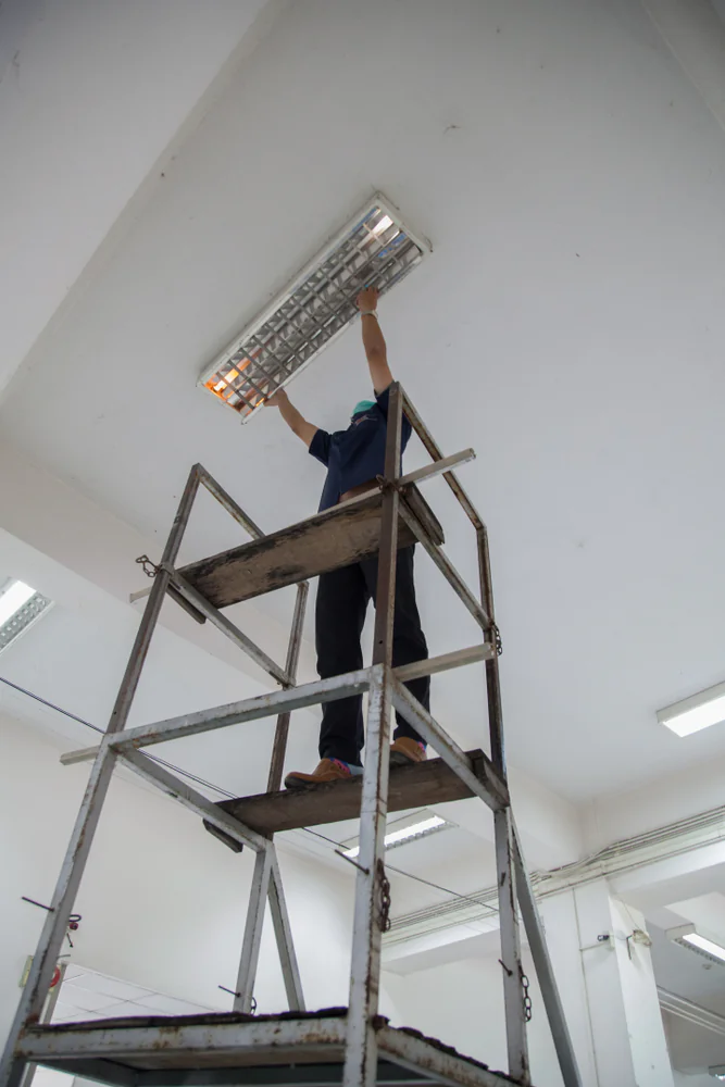 A person standing on top of a metal scaffold is changing or repairing a ceiling light fixture in a bright, white room.