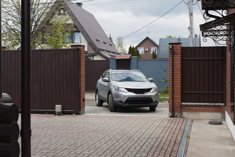 A silver SUV enters a driveway through an open brown gate, with brick pillars and residential houses visible in the background under a cloudy sky.