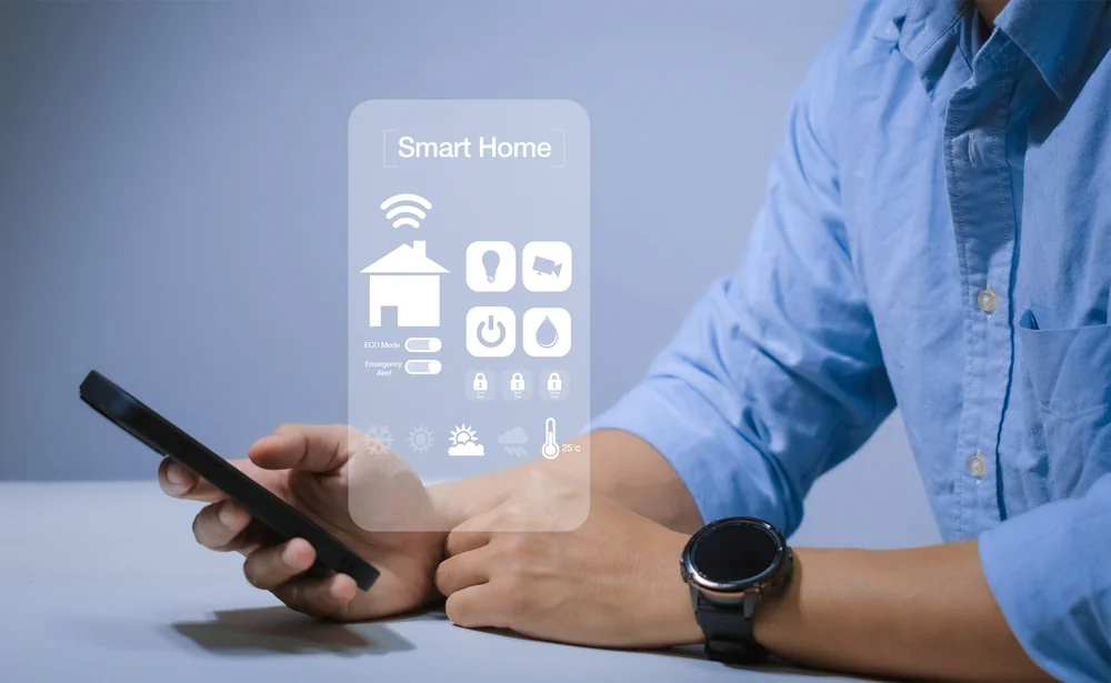 A person in a blue shirt uses a smartphone, with a transparent smart home interface displaying icons for Wi-Fi, lighting, security, climate control, and energy settings in front of them.