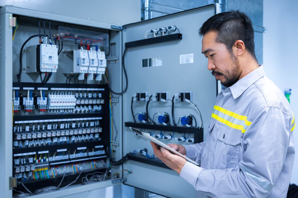 A technician in a gray uniform uses a tablet while inspecting and working on an open electrical control panel with numerous wires, switches, and components.