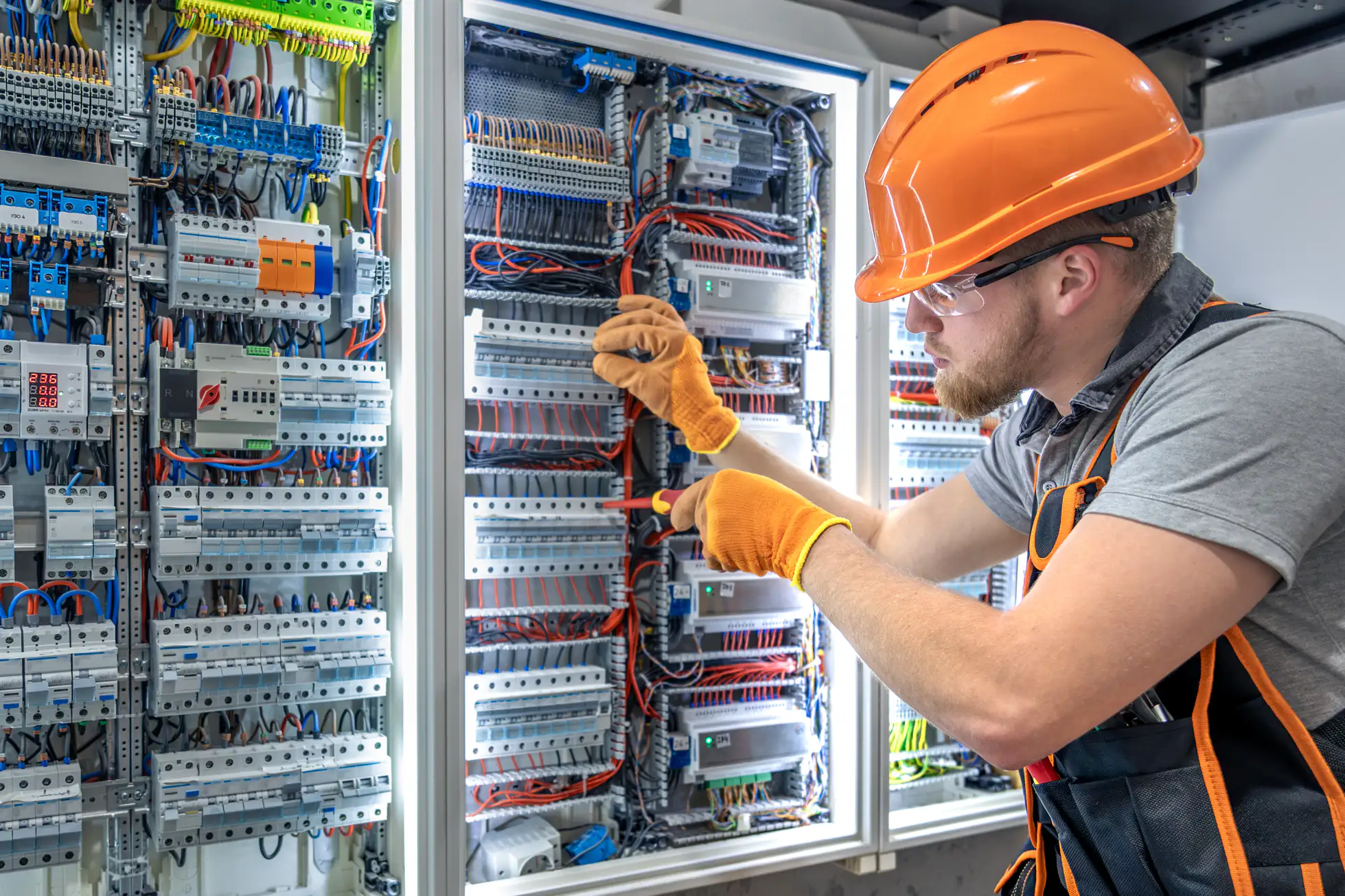 An electrician wearing a hard hat, safety glasses, and gloves uses a screwdriver to work on a large electrical control panel with many wires and components.