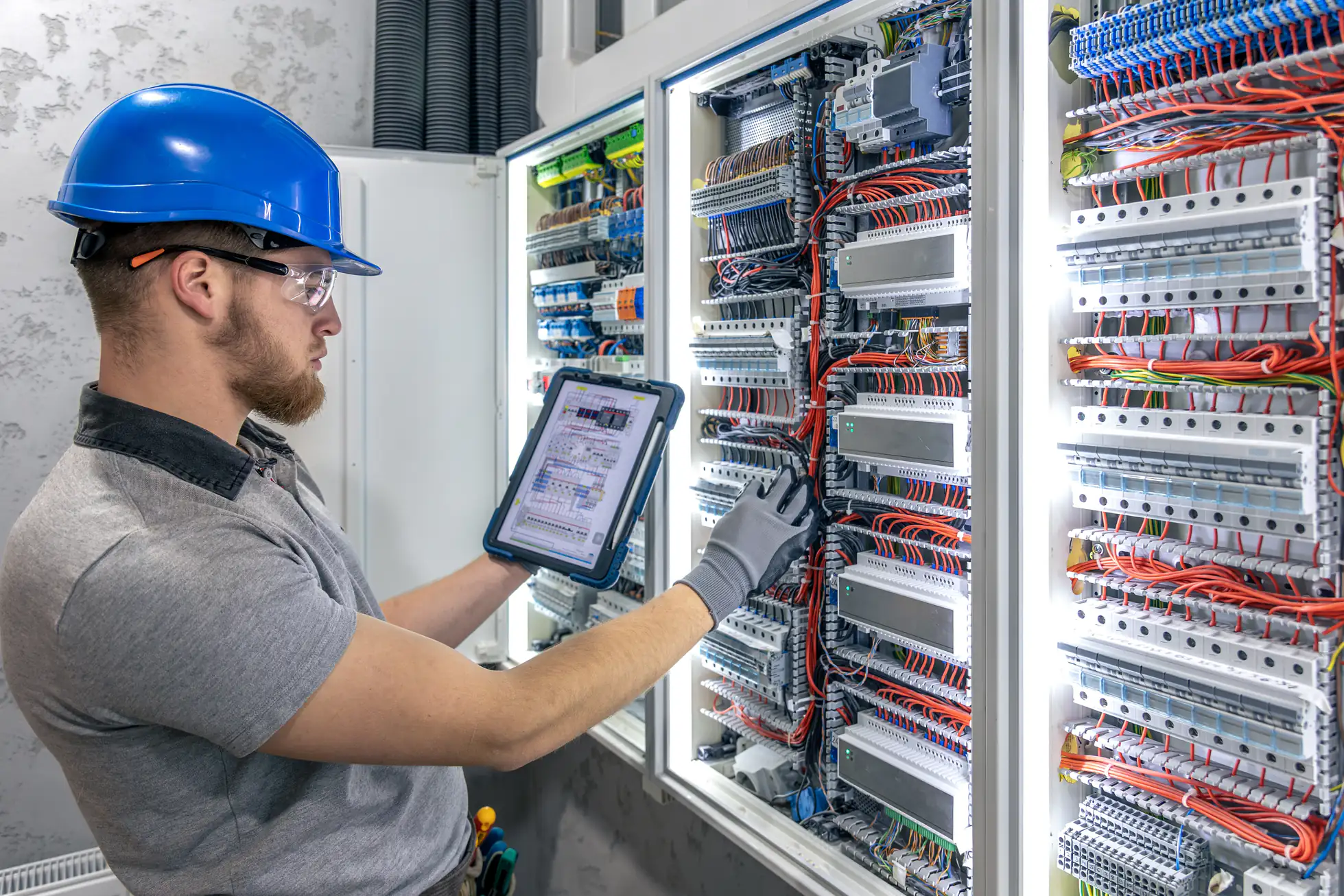 An electrician wearing a blue hard hat, gloves, and safety glasses inspects electrical control panels and wiring while holding a digital tablet in front of open equipment cabinets.
