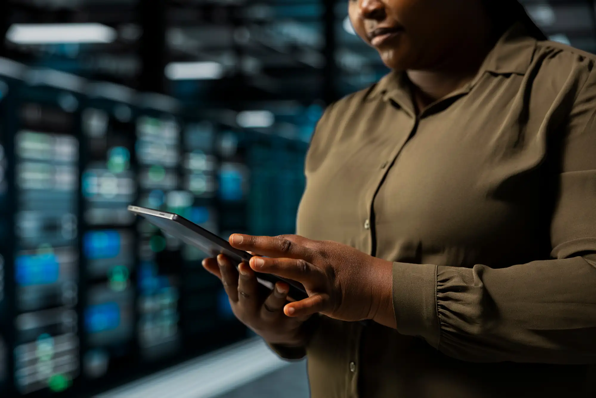 A person wearing an olive green shirt uses a tablet in a server room, with rows of blurred computer servers and equipment visible in the background.