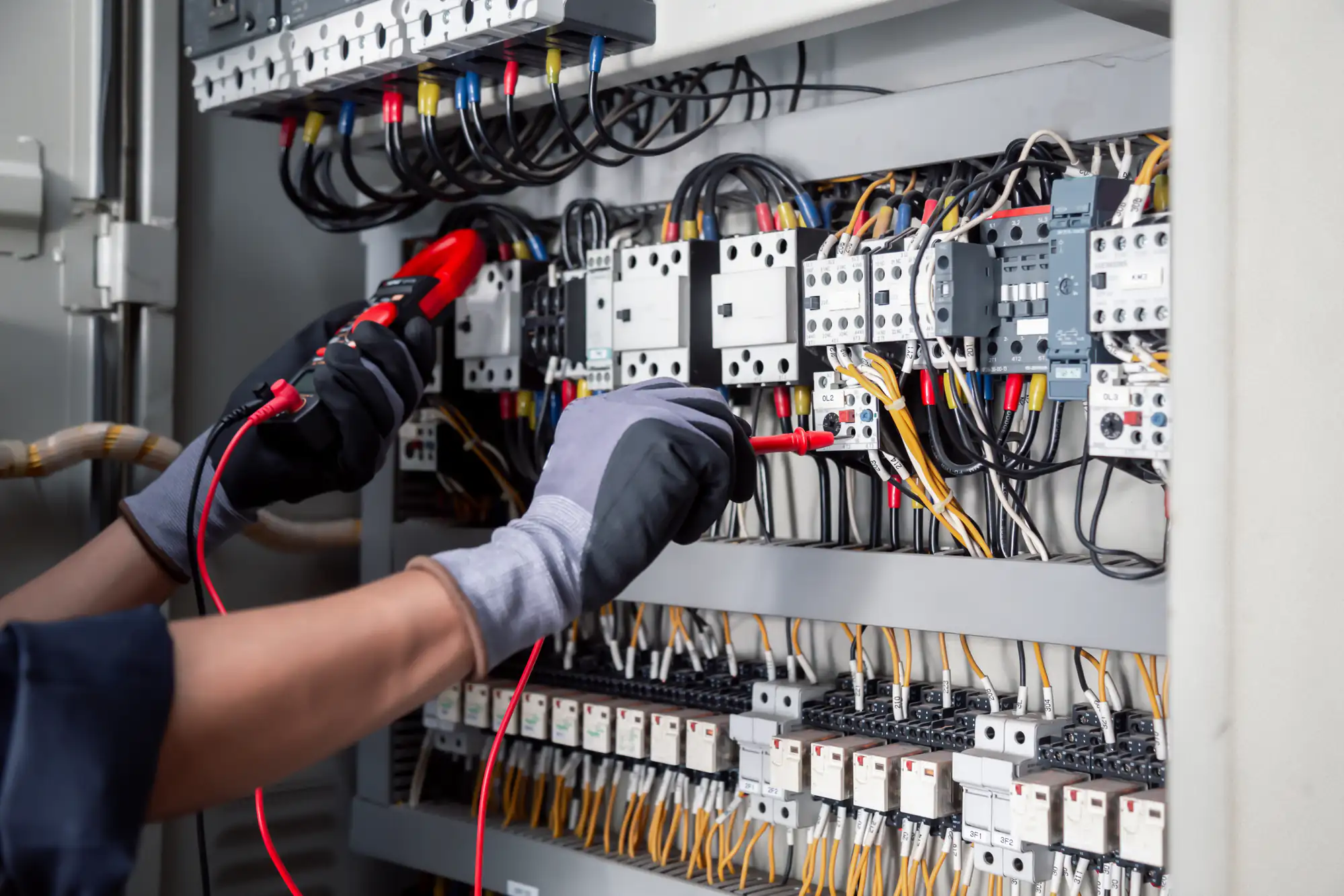An electrician wearing gloves uses a multimeter to test voltage in a control panel filled with wires, relays, and electrical components during electrical troubleshooting and repairs.