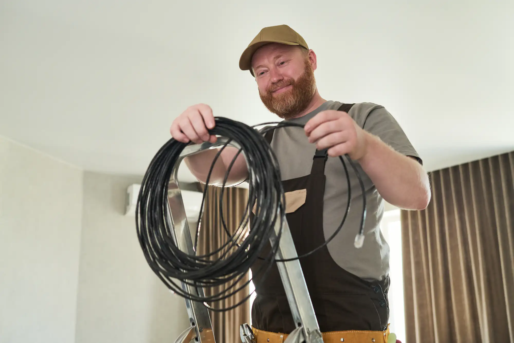 A smiling electrician in work overalls stands on a ladder, holding a coil of black cable indoors, ready for electrical troubleshooting and repairs, with brown curtains in the background.
