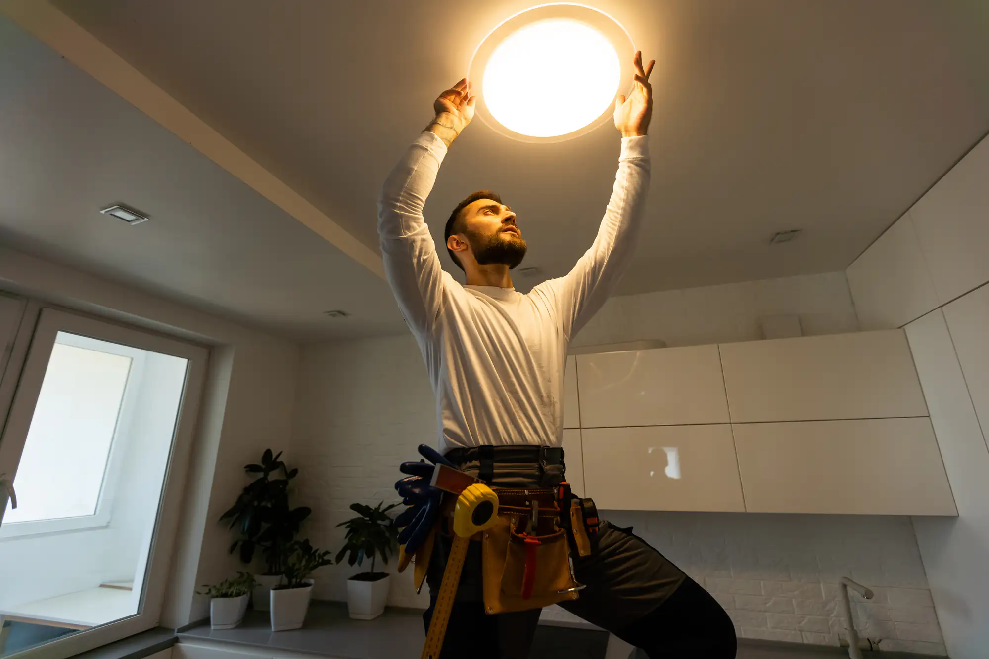 A man in a tool belt stands on a kitchen counter, reaching up to install or fix a ceiling light fixture in a modern, bright kitchen with white cabinets and potted plants.