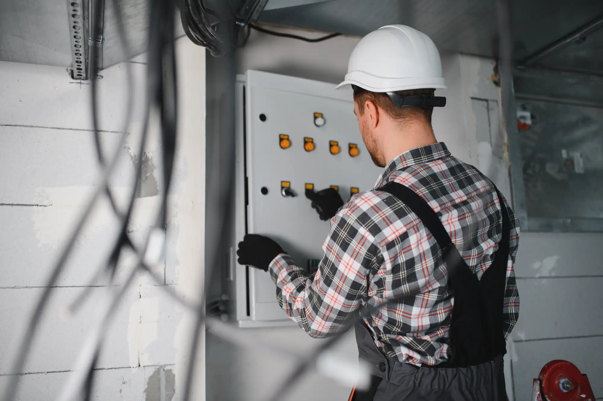 A person wearing a white hard hat, checked shirt, and overalls is working with an open electrical panel on a wall, adjusting switches or circuit breakers. Wires and construction materials are visible around them.