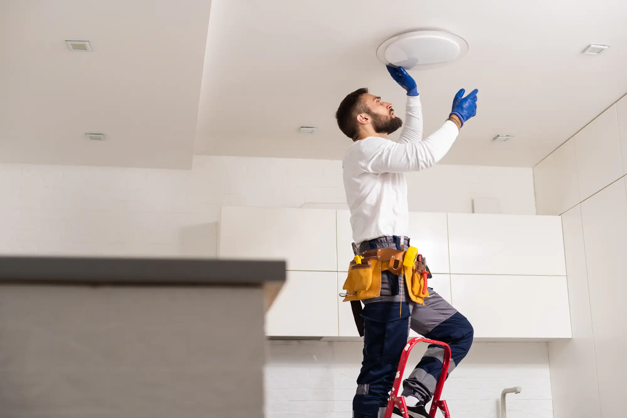 A man wearing work gloves and a tool belt stands on a step ladder, installing or fixing a ceiling light fixture in a modern kitchen.