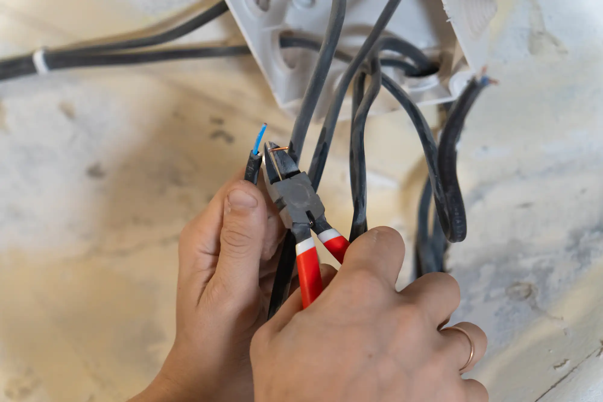 A person uses pliers to strip the insulation from an electrical wire among several black cables, preparing for electrical work. The hands and tools are in focus, with a junction box visible in the background.