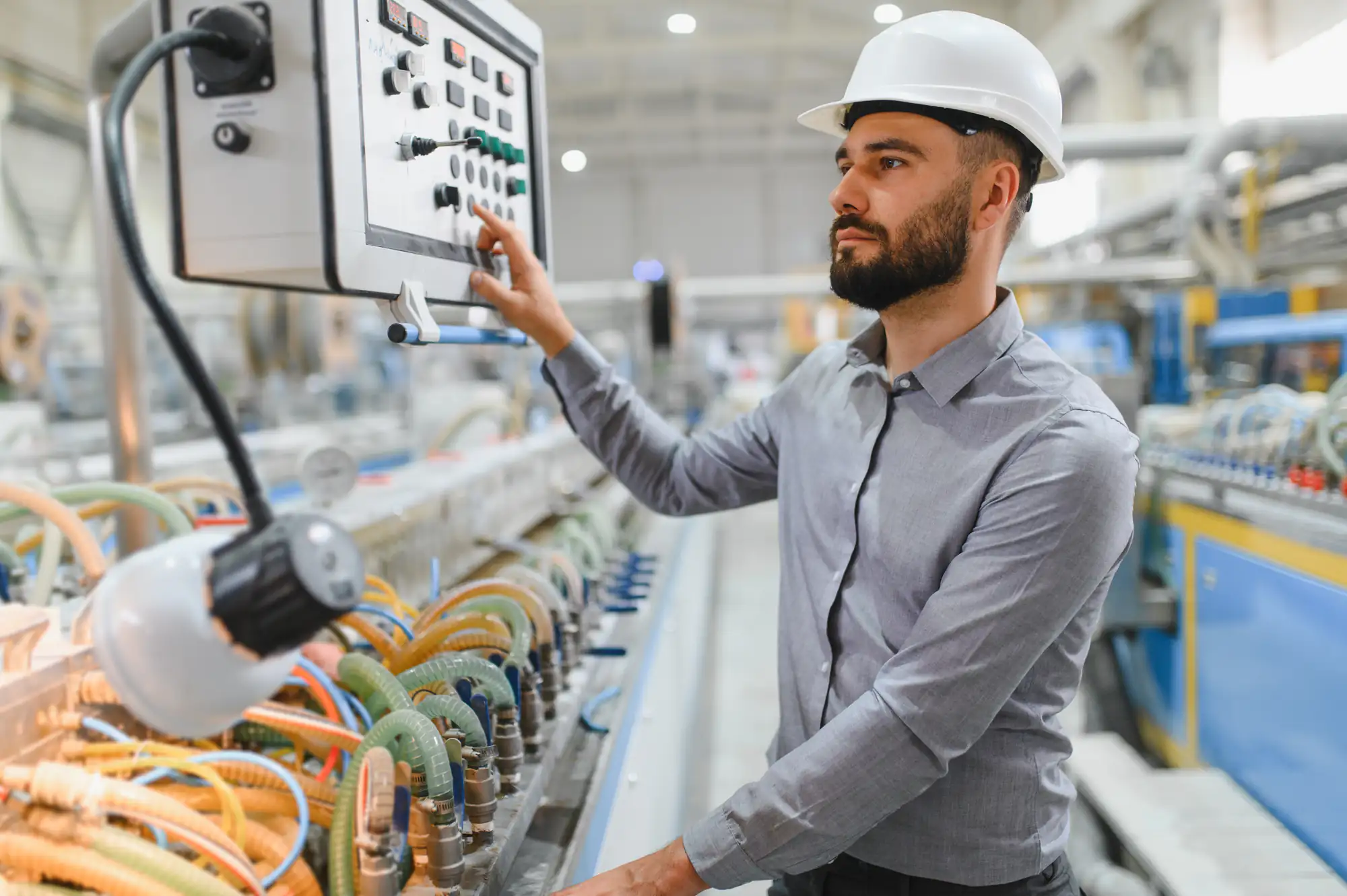 A man wearing a white hard hat and a gray shirt operates a control panel in an industrial factory, surrounded by machinery and colorful cables.