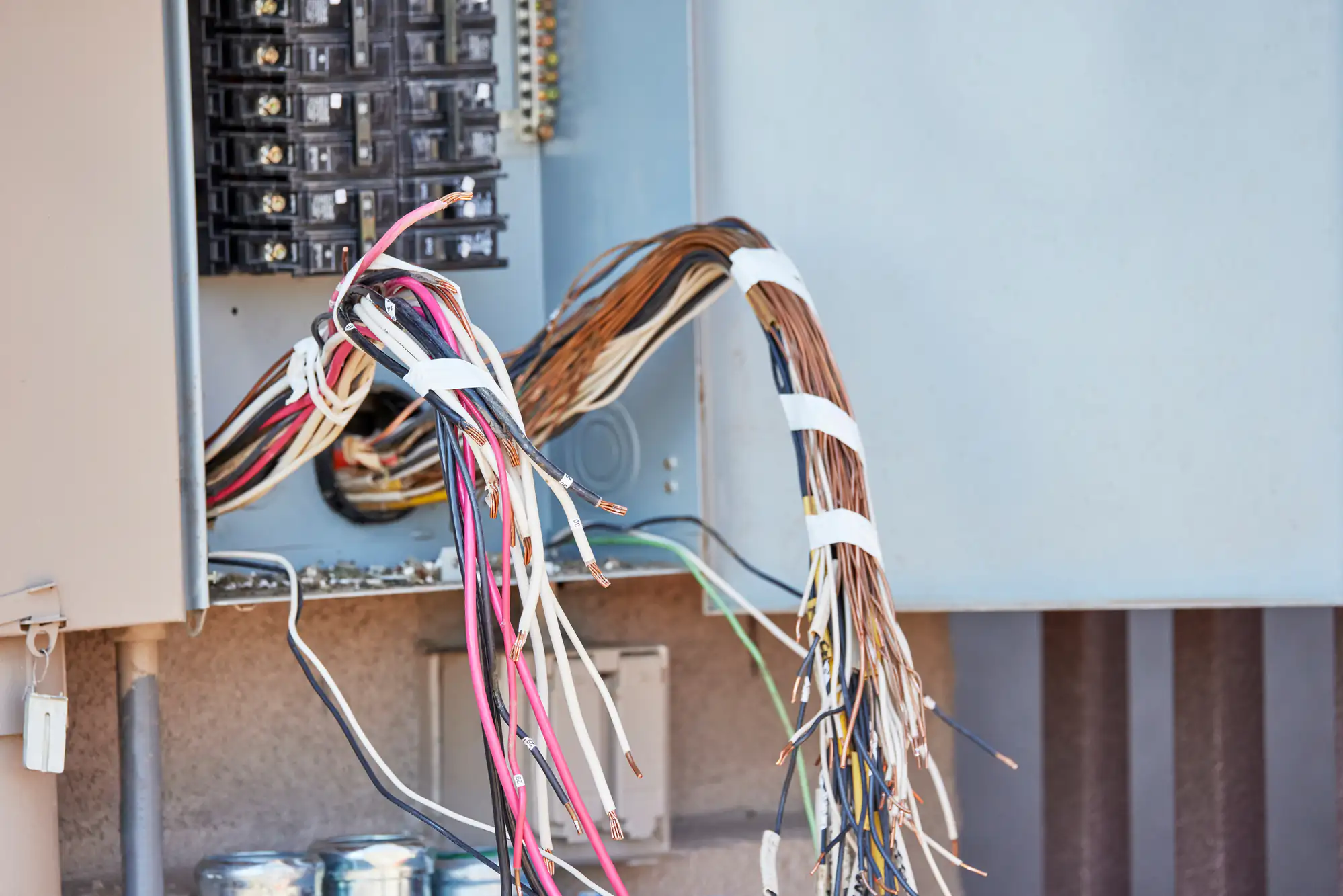A bundle of exposed electrical wires protrudes from an open electrical panel, with various colored wires secured by white zip ties. The panel's circuit breakers and metal components are visible.